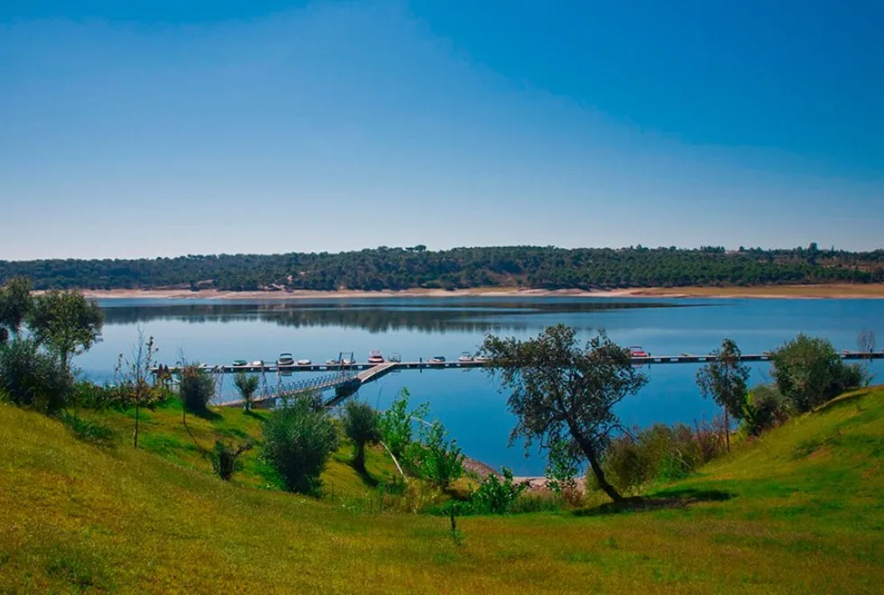 Vista panorâmica da Barragem de Montargil ao entardecer, com a água tranquila que reflete o céu e é rodeada por vegetação típica do Alentejo.
